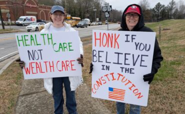 Demonstrators smile with their posters. One states "health care not wealth care" and the other says "Honk if you believe in the constitution."